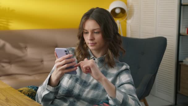 漂亮的年轻女子坐在扶手椅上 冷漠地浏览着手机 卷曲短发的女人在舒适的房间里舒适地呆在家里 高质量的4K镜头 — 图库视频影像
