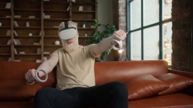 Hipster adam VR ya da sanal gerçeklik gözlüğü takıyor deri koltukta oturuyor ve evdeki oturma odasında video oyunu oynuyor. Teknoloji ve yenilikçi oyun kullanarak gündelik gömlekli genç adam.