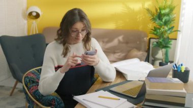 Gözlüklü sevimli bayan öğrencinin cep telefonuyla odada oturması hoş bir sürpriz oldu. Genç bayan güzel bir jest yapıyor, vücudunu heyecanla hareket ettiriyor. Yüksek kalite 4k görüntü