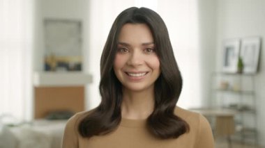 Smiling attractive young brunette woman model looking at camera. Happy beautiful 20s adult lady professional with pretty face and healthy smile posing alone indoors, slow motion close up portrait 4K