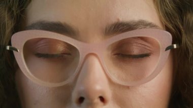 Close up beautiful light blue green eyes slow motion RED camera 4K shot. Happy young adult hipster gen z teen girl smiling face with curly hair wearing pink frame trendy eye glasses looking at camera