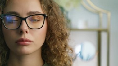 Portrait of attractive female student with curly brunette hair opening eyes and looking to camera in modern apartment. Close up face of beautiful woman in stylish glasses with black modern frame 4K