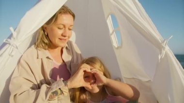 Aile bağlarını sevmek, gülmek, birlikte aşk kavramı. Sevecen, mutlu, genç bir kadın anne ve sevimli, küçük bir kız kalp şeklinde el hareketi yapıyorlar. Okyanus kıyısında altın gün batımında oturuyorlar.