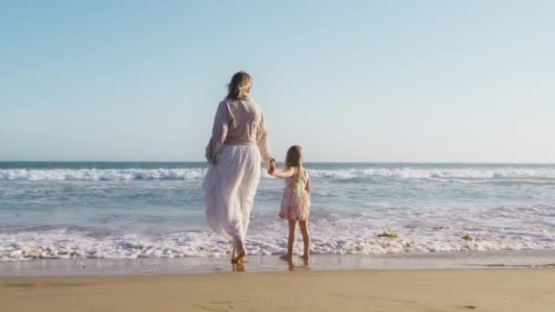 海に立っている母親と少女は 黄金の夕日の間の波を見ています 親と子供 幸せな子供時代 無料のライフスタイル 幸せな家族の概念 ママと娘のバックビュー一緒に手を保持 — ストック動画