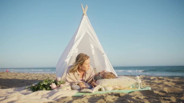 妈妈在孩子的帐篷里讲童话 妈妈睡觉前给小女儿读魔法故事 年轻的母亲躺在沙滩上白色帆布帐篷的毛毯上 和可爱的小女孩玩耍 — 图库视频影像