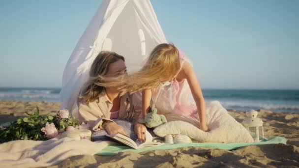 妈妈睡觉前给小女儿读魔法故事 年轻的母亲躺在沙滩上白色帆布帐篷的毛毯上 和可爱的小女孩玩耍 妈妈在童装帐篷里讲童话 — 图库视频影像