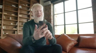 Happy blond haired middle age man laughing using smartphone looking at cell screen. Handsome hipster male take selfie watch games, make video call on mobile while sitting on leather sofa at loft home