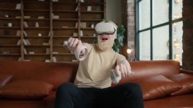 Teknoloji ve yenilikçi oyun kullanarak günlük gömlekli genç bir adam. Hipster adam VR ya da sanal gerçeklik gözlüğü takıyor deri koltukta oturuyor ve evdeki oturma odasında video oyunu oynuyor.