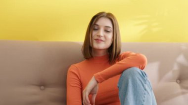 Evdeki kanepede oturan gülümseyen kadının portresi. Kamerada poz veren neşeli kız. Bayan öğrenci dersten sonra dinleniyor. Güzel bayan kafaya elle dokunuyor. Doğal makyajlı bir model, 4K görüntü.