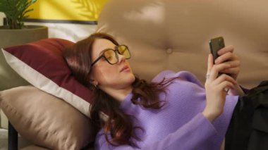 Akıllı telefonuyla kanepede uzanan neşeli kız. Telefondaki gülümseyen kadın. Evde cep telefonuyla konuşan olumlu bir bayan. Neşeli bayan telefon konuşmasından hoşlanıyor. İletişim konsepti