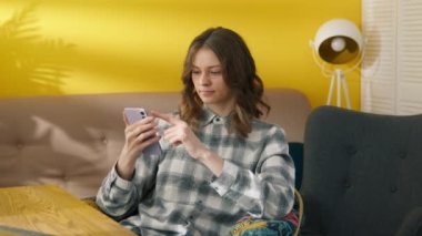 İnternetteki haberlere akıllı telefondan odaklanmış bir kadın. Evde gülen pozitif kız. Kolaj öğrencisi çevrimiçi dersler arasında dinleniyor. Cep telefonu kullanan gülümseyen kadın, 4K görüntü. 