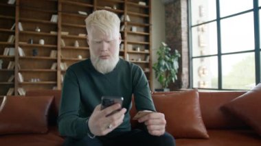 Hızlı ve kolay ödemeleri kullanan şaşırtıcı bir erkek müşteri. E-bankacılık uygulaması müşteri alımları online mağaza 4K. Kredi kartı ve akıllı telefon tutan etkileyici genç albino adam çatı katında kanepede oturuyor.