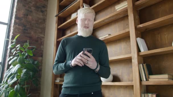 都市スタイルのアパートでスマートフォンを使用してロフトウィンドウにブロンドの髪を立てる中年のアルビノ男性のゆっくりとした動き ニューステキストメッセージをスクロールする男 テクノロジーライフスタイルスローモーションショット4K — ストック動画