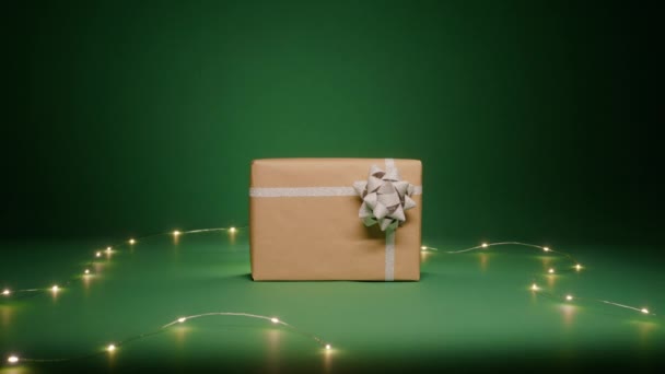 光沢のある銀の弓とクリスマスの軽いガーランドの装飾のクラフト紙のギフト用の箱が付いている魔法の夜の背景 緑の背景のクリスマスシーン スペースRedカメラをコピーします メリークリスマス 2024年 — ストック動画