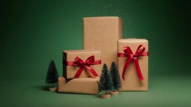 Altın hediye kutularının üzerine düşen kar taneleri zarif kırmızı fiyonklar ve yeşil arka planda izole çam ağaçları figürleri. Noel için sürpriz hediyeler. Mutlu 2024 Yeni Yıl sunumları
