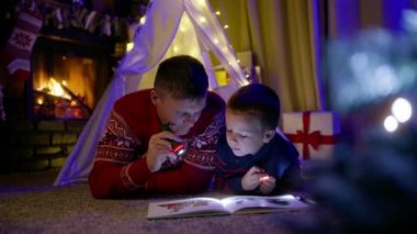 Baba ve oğul, oturma odasında el fenerleriyle oynuyor. Çadır bezinde Noel ağacı ve şömine üzerindeki çoraplar. Arka planda yanan şömine.