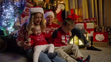 Mutlu aile Noel arifesinde Noel ağacının yanında toplandı. Kırmızı Noel Baba şapkalı genç ve güzel ebeveynler ve küçük kız bebek, anaokulu çocuğu şöminede eğleniyorlar.