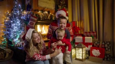 Mutlu bir aile, küçük bir kız çocuğu, anaokulu çocuğu ve kırmızı Noel Baba şapkalı güzel bir aile Noel arifesinde bir araya geldiler. Sıcak şömine RED 4K 'de birlikte vakit geçiriyorlar.