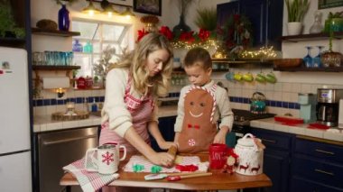 Çocuk annesine yardım ediyor, 4K numaralı raptiyeyle eğleniyor. Sarışın anne ve oğlu Noel temalı önlükler giyip, Noel arifesinde evde yemek için zencefilli kurabiye pişiriyorlar.