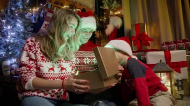 Üç kişilik beyaz bir aile Noel ağacının yanındaki şöminede oturuyor. Noel ruhu, birliktelik konsepti 4K görüntüsü. Sevimli küçük çocuk, parlak altın bir Noel hediyesi kutusuna bakıyor.