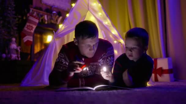两代家庭在一起度过的时光 在一个神奇的圣诞前夕 年轻的父亲带着他的小儿子躺在帆布帐篷里看书 书里有手电筒 — 图库视频影像