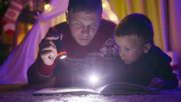 年轻的父亲和小儿子躺在用彩灯装饰的帐篷里 手握手电筒 大声朗诵童话故事 同时在壁炉边的小木屋里度过冬季的夜晚 — 图库视频影像