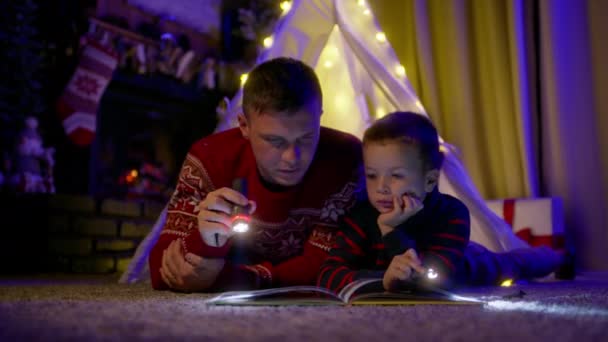 子供はクリスマスイブの魔法の話を注意深く聞いています 自宅のキャンバステントで息子と一緒に 懐中電灯のある若いハンサムな男は 本を強調し 小さな男の子に妖精の物語を読む — ストック動画