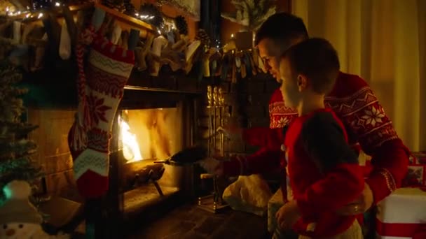 年轻的父亲和可爱的男孩用风箱把空气吹入壁炉 爸爸和儿子一起坐在舒适的壁炉边 旁边挂着长袜和圣诞花环灯 寒假和圣诞假期的家庭 — 图库视频影像