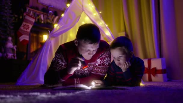 バックグラウンドで森を燃やしている居心地の良い暖炉の魔法のクリスマスイブにキャンバスのテントにフラッシュライト付きの本を読んでいる彼の赤ん坊の男の子と若い父親 2世代の家族が一緒に時間を過ごす — ストック動画