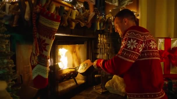 年轻英俊的男子穿着圣诞毛衣在舒适的壁炉旁的小木屋里 微笑的家伙用风箱把空气吹入火中 冬季和圣诞节假期的概念 慢镜头红外摄像机拍摄 — 图库视频影像