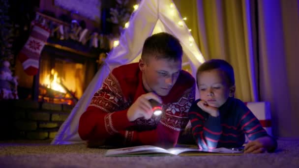 幸せな若い父と小さな息子は 明るく暖かい暖かい暖炉が背景に燃えている居心地の良いリビングルームに自宅でキャンバステントで本を読みました Redで撮影されたクリスマスの夜の魔法の居心地の良い雰囲気 — ストック動画