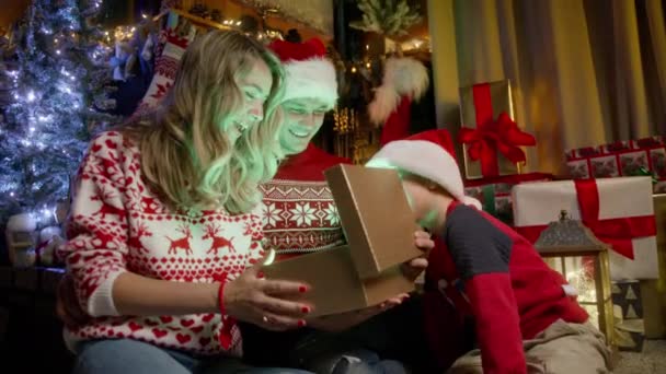 クリスマスツリーの近くの暖炉に座っている3人の白人家族 クリスマスの精神 トータルネスコンセプト4K映像は Redカメラを撮影しました 光沢のある黄金のクリスマスギフトボックスを見ている愛らしい少年 — ストック動画