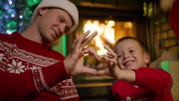 父親と息子の肖像画は 指で心臓の形を作ることに直面しています 家族が愛を分かち合い メリークリスマスと親戚を祝福し バックグラウンドで暖炉を燃やしました ハッピーホリデー — ストック動画