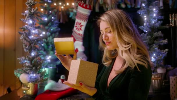 漂亮的年轻女子打开了一个金色的礼品盒 光芒照耀着她美丽的脸庞 迷人的微笑模特穿着天鹅绒翡翠裙 看起来很开心 背景4K的圣诞树灯 — 图库视频影像