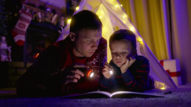 爸爸和儿子在家里的帆布帐篷里年轻英俊的男子 手电筒突出这本书和读童话故事给小男孩 圣诞节前夕 孩子们在听着神奇的故事 — 图库视频影像