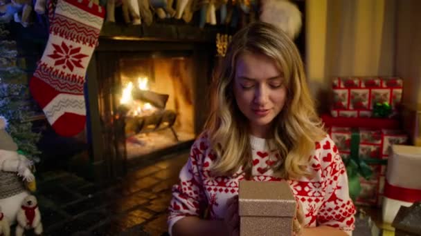魔法のクリスマスの夜4Kに居心地の良い装飾された暖炉に座っている白人美しい女性 幸せな魅力的なブロンドの女性が保持し 赤いカメラのための黄金の輝く輝きの輝く輝く輝きと光沢のある贈り物を示しています — ストック動画