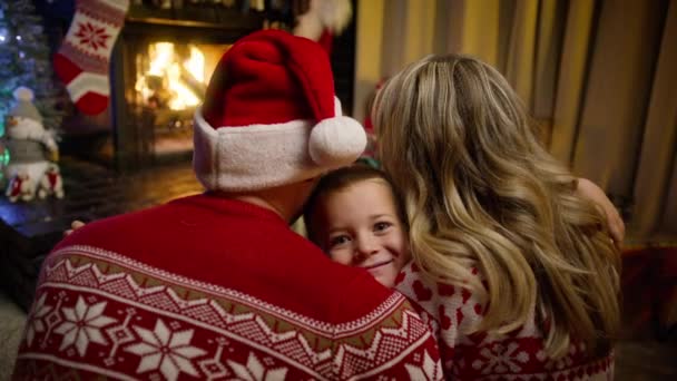 快乐的孩子拥抱他的父母 在装饰过的圣诞树前 可爱可爱的男孩抱着父母坐在壁炉前的画像 全家欢度圣诞快乐假期4K — 图库视频影像