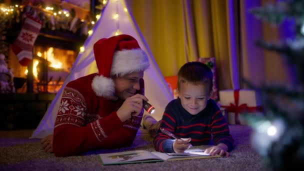 可爱有趣的小男孩许愿给圣诞老人写信 爸爸和孩子在一起玩4K游戏很开心 快乐的家庭爱爸爸 戴着圣诞红礼帽 带着手电筒和孩子在一起 — 图库视频影像