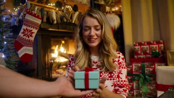 夫からの明るい青いティールボックスに存在することに興味を持っている若い30代の女性を微笑みます クリスマスプレゼントと赤いリボン箱に包まれた男性の手 装飾されたお祝いの暖炉に座っているブロンドの女性 — ストック動画