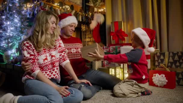 赤いサンタクロースの帽子のかわいい小さな笑顔の少年は クリスマスツリーの近くの暖炉に座っている若い両親にスタイリッシュな輝かしい黄金のクリスマスギフトボックスを与えます 冬の休日にプレゼントを持った幸せな子供 — ストック動画
