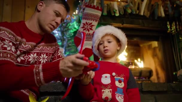 若い父親と興奮した子供がクリスマスプレゼントで遊んでいます かわいい家族の夢の自由飛行機の概念 笑顔の息子の夢は本当の幸せな休日を迎えました サンタハットで幸せな少年は飛行機のおもちゃで遊ぶ — ストック動画