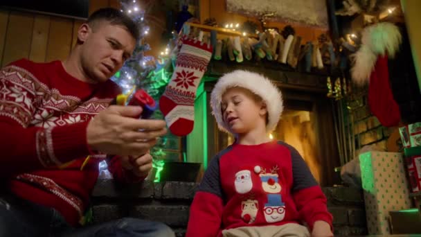 サンタハットの幸せな少年は飛行機のおもちゃで遊んでいます 若い父親と興奮した子供がクリスマスプレゼントで遊んでいます かわいい家族の夢の自由飛行機の概念 笑顔の息子の夢は本当の幸せな休日に来ました — ストック動画