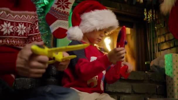 年轻的父亲和小孩戴着可爱的圣诞老人帽子 在壁炉边和五颜六色的飞机玩着有趣的游戏 圣诞前夜 快乐的男人 成年爸爸和小男孩玩玩具飞机 — 图库视频影像