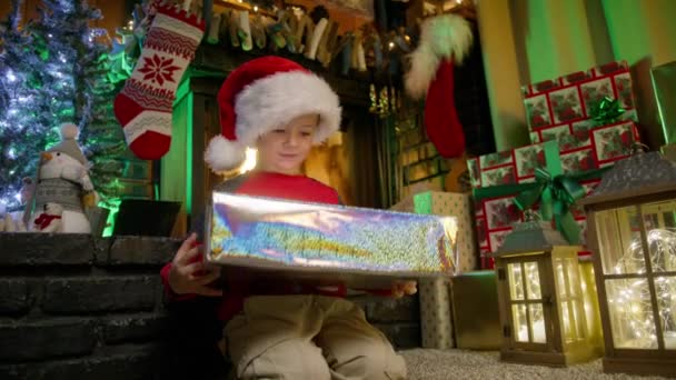 魔法のクリスマスの夜に輝くギフトボックスにプレゼントを開くことに興奮する幸せな小さな少年 赤いサンタの帽子の奇妙な子供はお祝いの休日を楽しんでいます 火の暖炉で家族と冬のお祝い — ストック動画