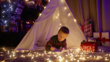 Anaokulu öğrencisi şöminenin yanında yatıyor ve Noel Baba 'ya kırmızı posta kutusundan Kuzey Kutbu' ndaki Noel Köyü 'ne göndermek için dilek listesi ve büyük hayalleriyle mektup yazıyor.