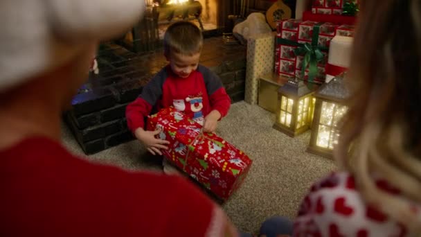 幸せな息子を見ている両親のリアビューは驚きで喜んでいます かわいい少年がクリスマスプレゼントを開けて 幸せに落ちる クリスマスイブ4Kで暖炉で子供と母と父親 — ストック動画