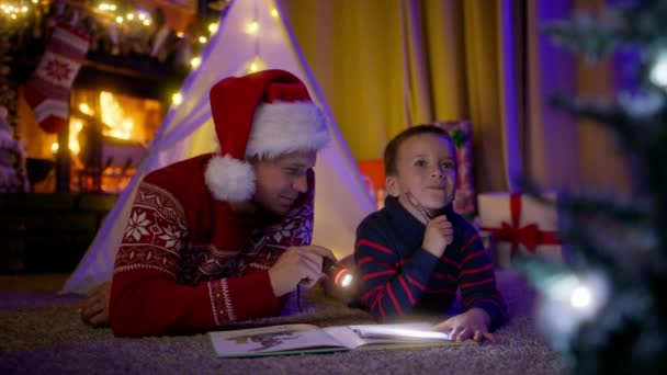 快乐的家庭爱戴着圣诞红礼帽的爸爸 手电筒看着多么可爱有趣的小儿子在向圣诞老人许愿和写信 爸爸和孩子在一起玩4K游戏很开心 — 图库视频影像