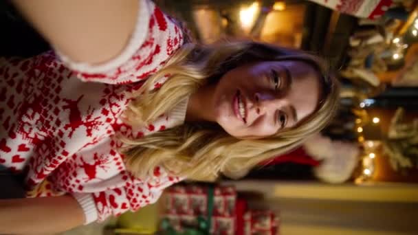 漂亮的金发女性穿着冬季毛衣 带着快乐的笑容 在智能手机上享受着视频通话 美丽的女孩在圣诞树前与网络摄像头聊天 还在彩灯下拍摄了4K的垂直镜头 — 图库视频影像