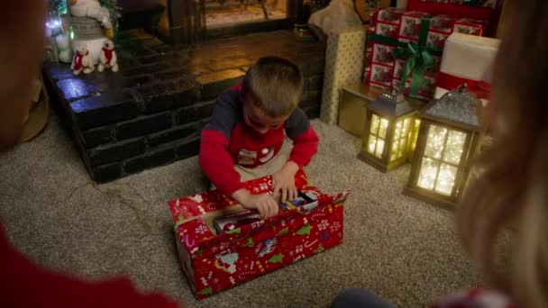 カリフォルニア州ビッグベアキャビン 2022年12月 幸せな愛らしい息子を見ている両親のリアビューは驚きで喜んでいます クリスマスイブ赤いカメラで幸せでクリスマスプレゼントを開くかわいい男の子 — ストック動画