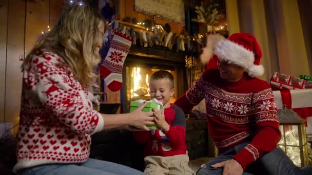 非常に幸せな小さな少年は 装飾されたクリスマスツリーの近くで驚きのプレゼントを得ることに興奮しています 暖炉に座っている幸せな家族 かわいいキッズラッピングギフトボックス シネマティッククリスマスイブレッドカメラ — ストック動画